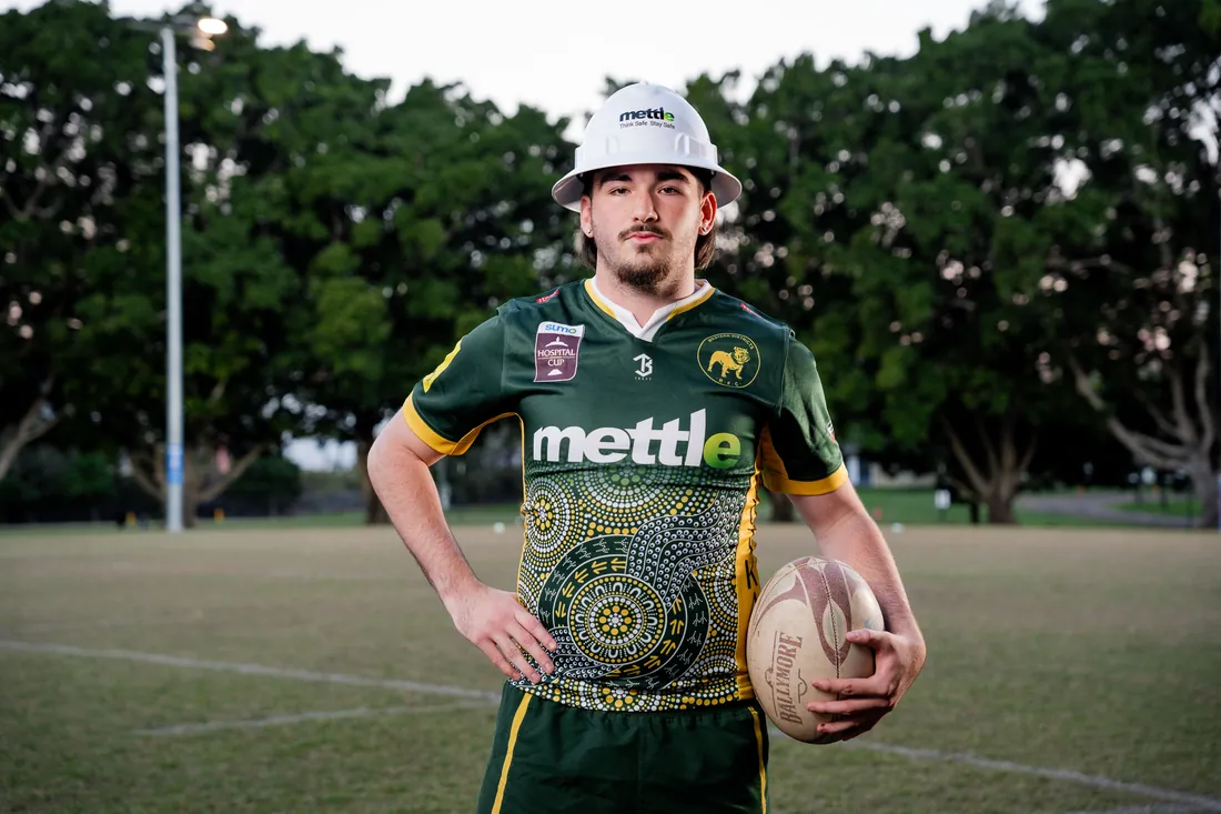 Young player wearing a Mettle jersey with Aboriginal artwork and a Mettle hard hat, holding a rugby ball on the field